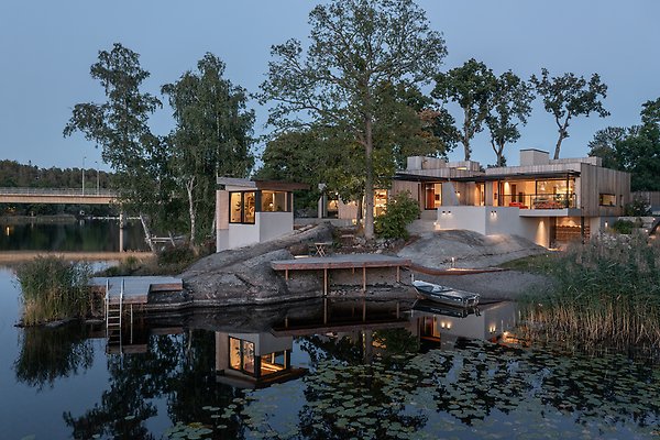 Ett modernt hus vid en sjö, byggt på klippor och omgivet av träd.